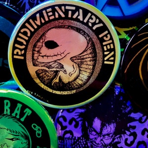 May include: A collection of colorful round pins with various graphic designs. One pin features a black and white illustration of a skull with the text "Rudimentary Pen" in a yellow circle. Another pin features a green and black illustration of a person with the text "Rat Noise" in a yellow circle. A third pin features a purple and black illustration of a creature with a menacing expression.