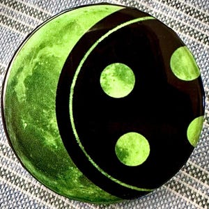 May include: A round ceramic plate with a green and black design. The plate features a green crescent shape and a black section with green polka dots. The plate has a glossy finish. The design is reminiscent of a yin-yang symbol.