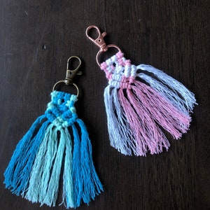 Puede incluir: Dos llaveros de macramé hechos a mano con borlas. Un llavero es azul y verde, el otro es rosa y blanco.