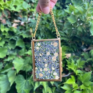 May include: A rectangular, gold-coloured pendant with a chain, filled with dried lavender and small white flowers. The pendant is held up against a backdrop of green foliage.