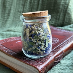 May include: A small, clear glass jar with a cork stopper, filled with a mixture of dried blue flower petals, small white star-shaped blossoms, and green leaves. The jar is resting on a brown leather-bound book with embossed floral patterns.