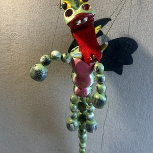 Puede incluir: Un dragón de madera verde con lengua roja y alas negras. El dragón tiene una cuerda atada a la cabeza y una serie de cuentas de madera para el cuerpo.