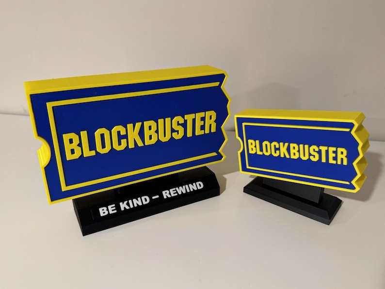 以下が含まれることがあります： 青と黄色のブロックバスターのチケット型のサインが2つ。大きなサインには、ベースに「BE KIND - REWIND」の文字があります。