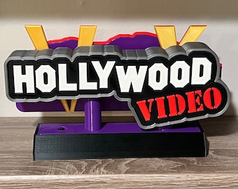 以下が含まれることがあります： 「HOLLYWOOD VIDEO」と書いてある、紫と黒の3Dプリントされた看板。側面には黄色の稲妻が描かれている。