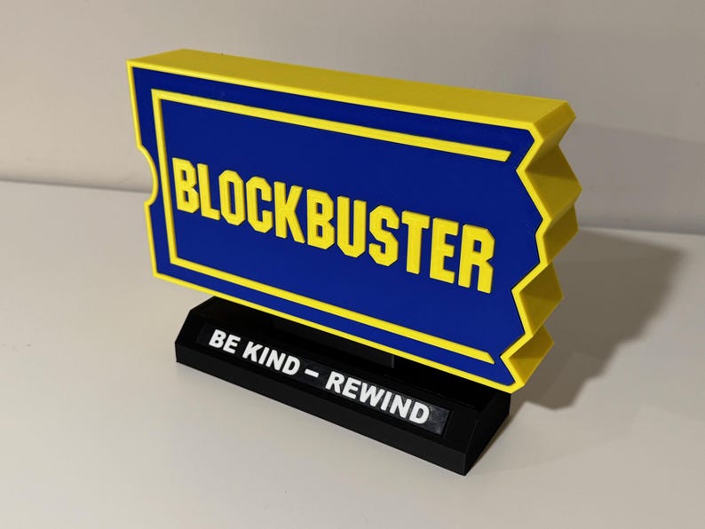 以下が含まれることがあります： 黒い台座に「BE KIND - REWIND」の文字が書かれた黄色と青のブロックバスタービデオ店の看板です。
