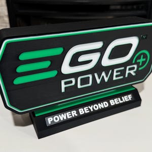 May include: A black and green sign with the text "EGO POWER" in white. The sign has a green border and a green plus symbol. The base of the sign reads "POWER BEYOND BELIEF" in white text.