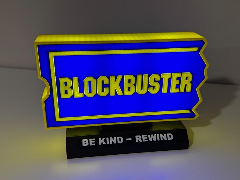 以下が含まれることがあります： 黒い台座に「BE KIND - REWIND」の文字が書かれた黄色と青のブロックバスターの看板です。