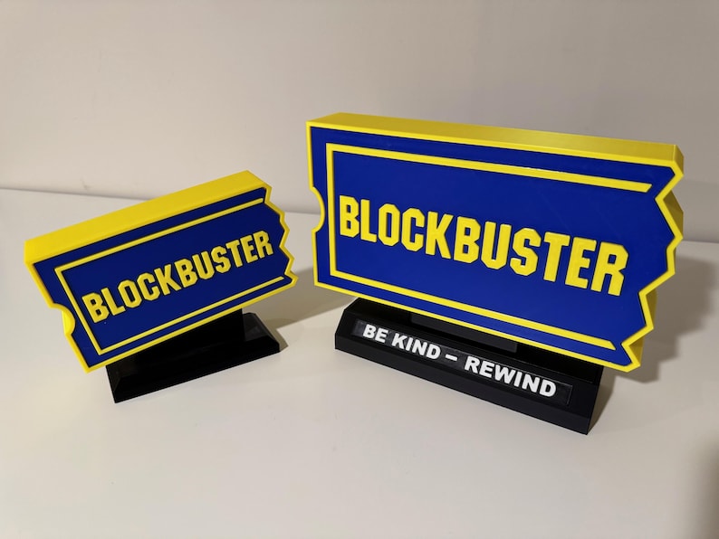 以下が含まれることがあります： 黄色と青のブロックバスターのチケットの形をしたサインが2つ。大きなサインには、ベースに「BE KIND - REWIND」という文字があります。