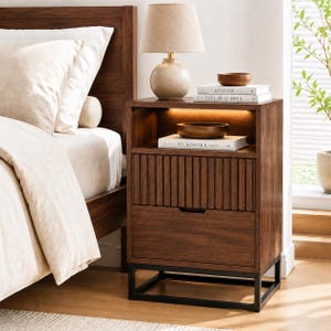 Peut inclure: Une table de chevet en bois foncé avec une base en métal noir, comprenant une étagère ouverte avec éclairage intégré, un tiroir à façade cannelée et un tiroir lisse. Une lampe beige et deux livres intitulés Kinfolk et Live Beautiful sont posés dessus.