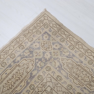 Puede incluir: Primer plano de una secci&oacute;n de una alfombra beige y azul estampada. La alfombra presenta un dise&ntilde;o floral y geom&eacute;trico detallado con un borde. La alfombra est&aacute; colocada sobre una superficie blanca.