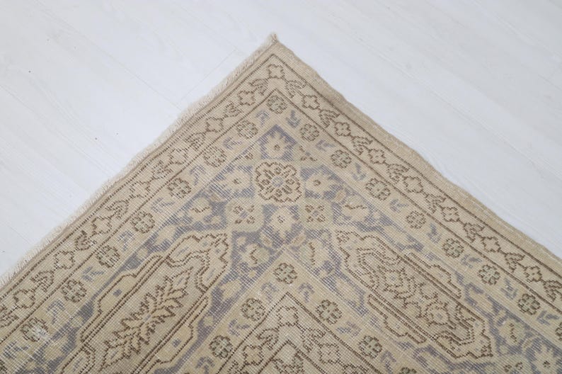 Puede incluir: Primer plano de una secci&oacute;n de una alfombra beige y azul estampada. La alfombra presenta un dise&ntilde;o floral y geom&eacute;trico detallado con un borde. La alfombra est&aacute; colocada sobre una superficie blanca.