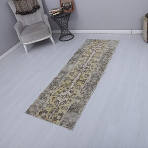 May include: A long, narrow rug with a distressed appearance, featuring a central floral design in yellow, grey, and beige. The rug is suitable for a hallway or entryway. It is placed on a light-coloured floor.
