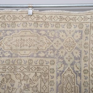 Puede incluir: Fragmento de una alfombra vintage con una combinaci&oacute;n de colores beige y lila claro. La alfombra presenta intrincados patrones florales y geom&eacute;tricos en tonos marr&oacute;n y beige. La alfombra est&aacute; colgada de una barra met&aacute;lica con clips.
