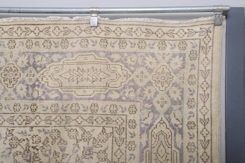 Puede incluir: Fragmento de una alfombra vintage con una combinaci&oacute;n de colores beige y lila claro. La alfombra presenta intrincados patrones florales y geom&eacute;tricos en tonos marr&oacute;n y beige. La alfombra est&aacute; colgada de una barra met&aacute;lica con clips.
