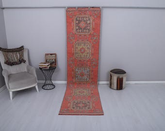 Puede incluir: Una alfombra larga y rectangular de color rojo con un patrón repetitivo de medallones cuadrados en tonos marrones, beige y verdes. La alfombra se exhibe verticalmente contra una pared gris, mostrando su intrincado diseño y su cálida paleta de colores.