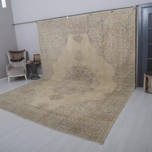 Puede incluir: Una alfombra grande, beige y marr&oacute;n con un dise&ntilde;o central ornamentado. La alfombra est&aacute; colocada sobre un suelo de color claro, con una parte drapeada sobre una barra de metal. Los intrincados detalles y los tonos neutros sugieren un estilo vintage o tradicional.