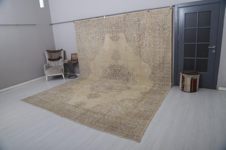 Puede incluir: Una alfombra grande, beige y marr&oacute;n con un dise&ntilde;o central ornamentado. La alfombra est&aacute; colocada sobre un suelo de color claro, con una parte drapeada sobre una barra de metal. Los intrincados detalles y los tonos neutros sugieren un estilo vintage o tradicional.
