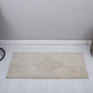 Op de afbeelding: Een rechthoekig, beige tapijt met een centraal patroon. Het tapijt ligt op een witte vloer, met een zwarte bijzettafel en een poef met patroon op de achtergrond. Het tapijt meet ongeveer 60 x 120 cm.
