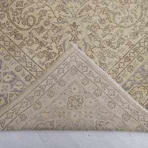 Puede incluir: Alfombra vintage beige y gris con intrincados patrones florales y geom&eacute;tricos. Una esquina de la alfombra est&aacute; doblada, revelando la parte inferior. El dise&ntilde;o presenta un medall&oacute;n central y detalles de borde, creando una est&eacute;tica cl&aacute;sica y envejecida.
