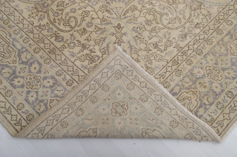 Puede incluir: Alfombra vintage beige y gris con intrincados patrones florales y geom&eacute;tricos. Una esquina de la alfombra est&aacute; doblada, revelando la parte inferior. El dise&ntilde;o presenta un medall&oacute;n central y detalles de borde, creando una est&eacute;tica cl&aacute;sica y envejecida.