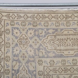 Puede incluir: Primer plano de una alfombra vintage con una paleta de colores crema y azul claro. La alfombra presenta un intrincado patr&oacute;n floral y geom&eacute;trico con acentos marrones. Cuelga de una barra de metal con clips.