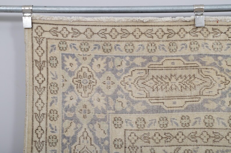 Puede incluir: Primer plano de una alfombra vintage con una paleta de colores crema y azul claro. La alfombra presenta un intrincado patr&oacute;n floral y geom&eacute;trico con acentos marrones. Cuelga de una barra de metal con clips.