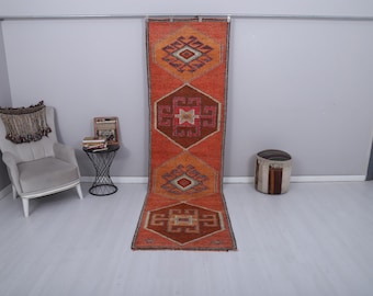 Könnte beinhalten: Ein langer, rechteckiger, orangefarbener Teppich mit geometrischen Mustern in Braun-, Orange- und Cremetönen. Der Teppich liegt auf einem hellgrauen Boden, mit einer grauen Wand im Hintergrund. Das Design des Teppichs zeigt sich wiederholende Diamanten und geometrische Formen.