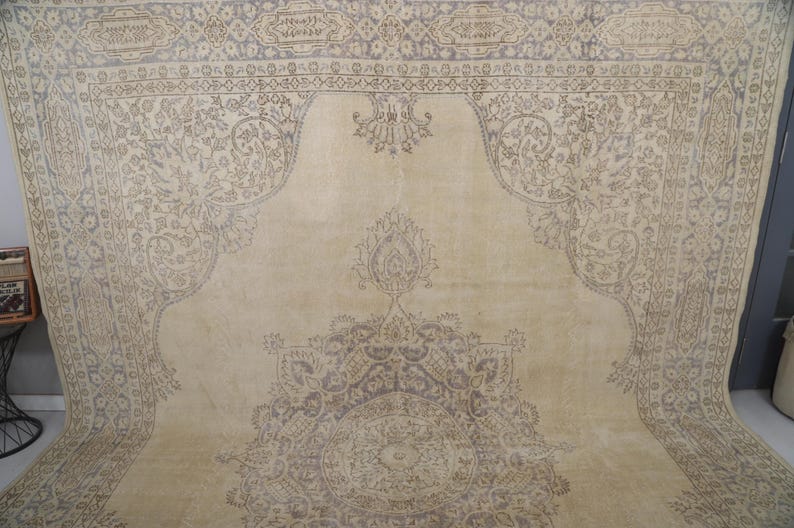 Puede incluir: Una alfombra beige grande con un intrincado patr&oacute;n floral descolorido en tonos grises y topo. La alfombra presenta un dise&ntilde;o de medall&oacute;n central y bordes ornamentados, creando una est&eacute;tica vintage. La alfombra est&aacute; doblada, mostrando su tama&ntilde;o.