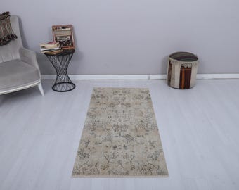 May include: A rectangular beige rug with a floral pattern in shades of brown and gray. The rug is placed on a light-colored wooden floor. A gray armchair, a black side table, and a patterned ottoman are also in the room.