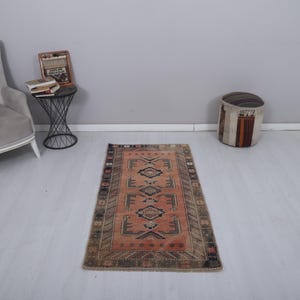 May include: A rectangular, vintage-style rug with a central geometric pattern in shades of peach, brown, and black. The rug has a dark border and is placed on a light-colored floor. The rug's dimensions are approximately 3 x 5 feet.