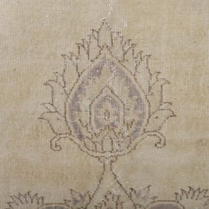 Puede incluir: Primer plano de una alfombra beige con un dise&ntilde;o floral central en tonos morados y marrones apagados. La alfombra tiene un aspecto envejecido y vintage, con signos de desgaste, lo que sugiere edad y uso. El patr&oacute;n es sim&eacute;trico.