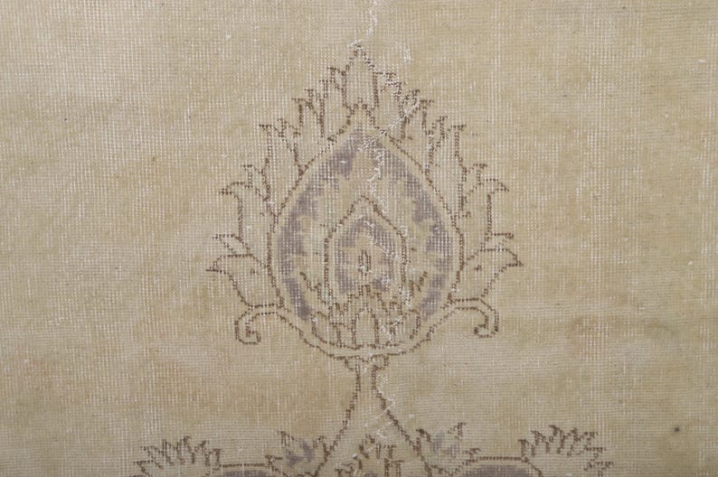 Puede incluir: Primer plano de una alfombra beige con un dise&ntilde;o floral central en tonos morados y marrones apagados. La alfombra tiene un aspecto envejecido y vintage, con signos de desgaste, lo que sugiere edad y uso. El patr&oacute;n es sim&eacute;trico.