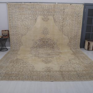 Puede incluir: Una alfombra grande, beige y marr&oacute;n con un dise&ntilde;o de medall&oacute;n central ornamentado. La alfombra se exhibe sobre un suelo de color claro, con una silla, una mesa auxiliar y una puerta visibles en el fondo. Las dimensiones de la alfombra no son visibles.