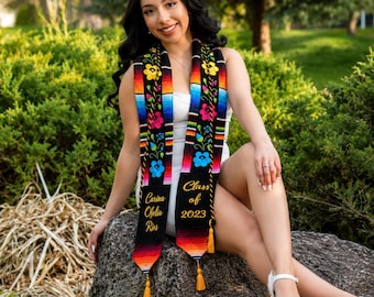 May include: A graduation stole with colorful floral embroidery and the text "Carina Ofelia Rios" and "Class of 2023." The stole has a black base with vibrant stripes and gold tassels. The stole is worn over a white outfit.