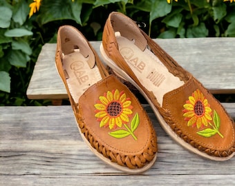 Könnte beinhalten: Ein Paar braune Ledersandalen mit gestickten Sonnenblumen. Die Sandalen haben ein geflochtenes Riemendetail und der Markenname "JAB" ist auf der Innenseite der Sandalen sichtbar.