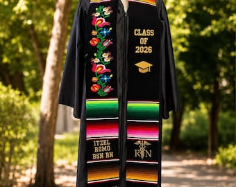 Puede incluir: Una toga de graduación negra con una colorida banda bordada. La banda presenta diseños florales, el texto "CLASS OF 2026" y el nombre "ITZEL ROMO BSN RN". La banda también tiene un símbolo médico.