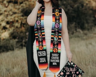 May include: A woman wearing a white dress and a colorful graduation stole with the text "CSUSB 2024" and "YARELI CHAVEZ SANCHEZ MPA". She is holding a graduation cap with the text "What like it's hard?" in pink and black lettering.