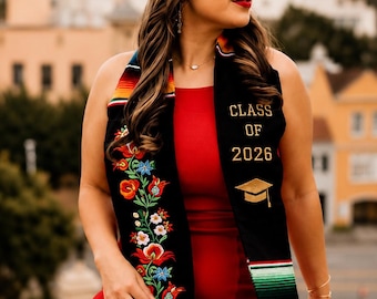 May include: A woman wearing a black graduation cap and a red dress. She is wearing a black sash with the text "CLASS OF 2026" and a floral design. The sash also has the text "FIRST GENERATION".