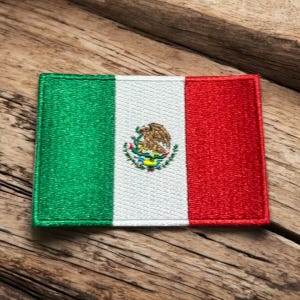 May include: A green, white and red embroidered patch with the Mexican coat of arms in the centre. The patch is on a wooden surface.