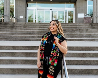 May include: A person wearing a black top and pants, with a colorful graduation sash that reads "RDH BSDH 2024." The sash features floral and geometric patterns. The background is a library with the text "JOHN F. KENNEDY LIBRARY."