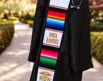 Peut inclure: Une toge de remise de diplôme noire portée avec une étole serape colorée à rayures horizontales rouges, blanches, vertes et bleues. L'étole est brodée du nom SONIA RAMIREZ et d'un symbole caducée doré au-dessus des lettres RN.