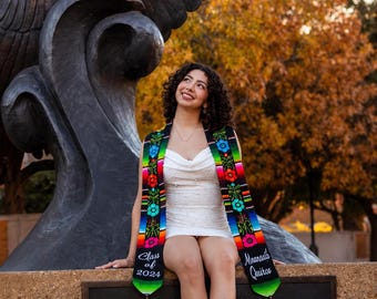 May include: A woman wearing a white dress and a colorful graduation stole with the text "Class of 2024" and "Mnandalis Quiros". The stole has a black background with floral and geometric patterns. She is sitting in front of a bronze statue.