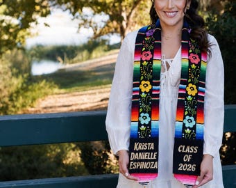 May include: A black graduation stole featuring a vibrant rainbow-striped pattern, embroidered colorful floral designs, and gold lettering that reads Krista Danielle Espinoza and Class of 2026, finished with gold tassels at the bottom ends.