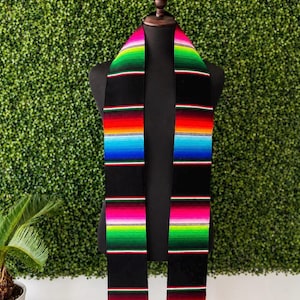 Könnte beinhalten: Ein schwarzer Serape-Schal mit leuchtenden Streifen in Rot, Orange, Grün, Blau und Pink. Die Enden des Schals haben weiße Fransen. Der Schal wird auf einer schwarzen Schaufensterpuppe vor einem grünen Hintergrund präsentiert.