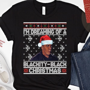 Op de afbeelding: Zwarte kersttrui met een zwart-wit sneeuwvlokkenpatroon. De trui heeft een portret van een man met een kerstmanmuts en de tekst "I'm Dreaming of a Blackity-Black Christmas".