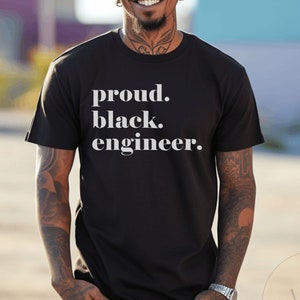 May include: Black crew neck t-shirt with the text "proud. black. engineer." in white. The shirt is worn by a person with tattoos and a watch. The background is blurred with shades of blue and purple.