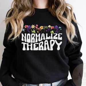May include: Black sweatshirt with a white graphic that says "Normalize Therapy" in a retro font. The graphic is surrounded by a row of colourful flowers.