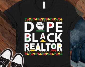 May include: Black t-shirt with the words "DOPE BLACK REALTOR" in white, red, and green. The design includes a raised fist and geometric patterns in red, yellow, and green. The shirt is displayed on a wooden surface with jeans and sneakers.