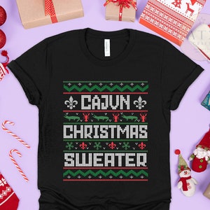 May include: Black t-shirt with a green, red, and white Christmas sweater design. The design features a fleur-de-lis, alligators, and crawfish. The text on the shirt reads "Cajun Christmas Sweater".