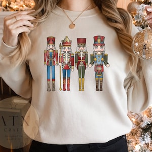 May include: A white sweatshirt with a graphic of four nutcracker soldiers. The nutcrackers are wearing colorful uniforms and holding various instruments. The text "ATX CRAFT designed in austin, tx" is printed on the bottom left corner of the sweatshirt.